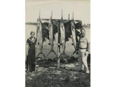 Vintage photo of man and woman standing next to a row of hanging swordfish
